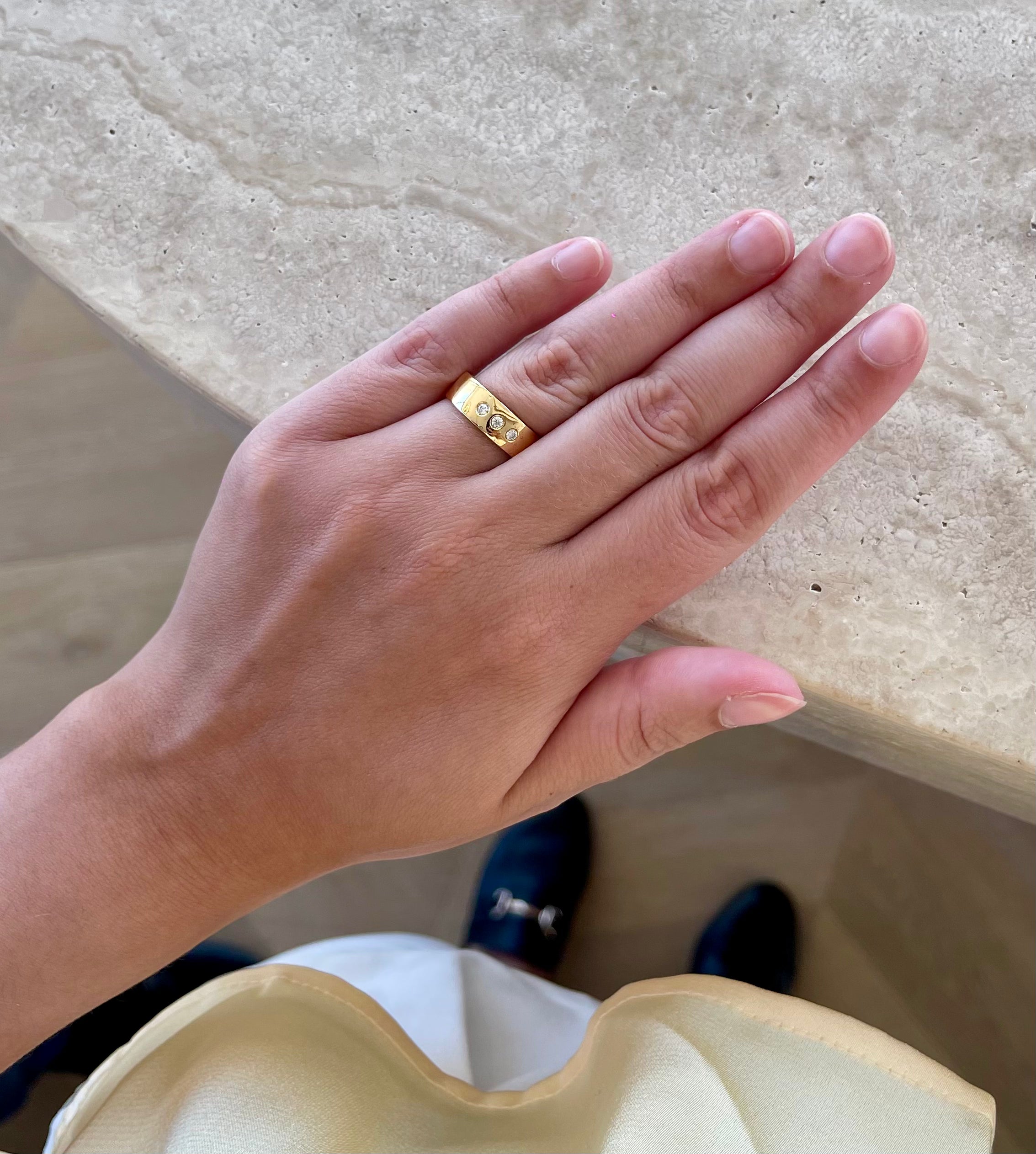 Model wearing DOLCE 6mm ring in yellow gold next to a beige marble bar wearing black loafers from STEVE MADDEN, a yellow summer top and a white corporate short.